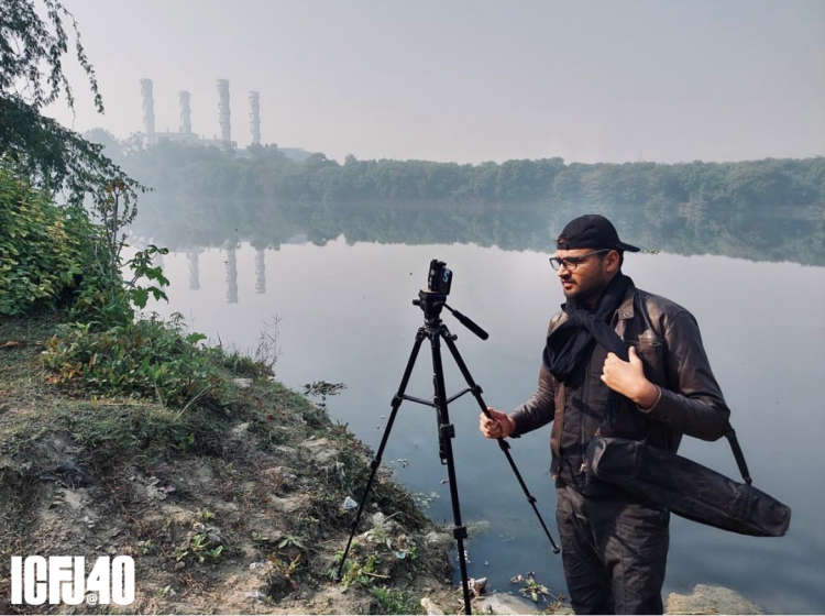 Rohit Upadhyay stands in the foreground holding a tripod. He's standing on the shore of a large body of water, with shrubbery and water in the background.