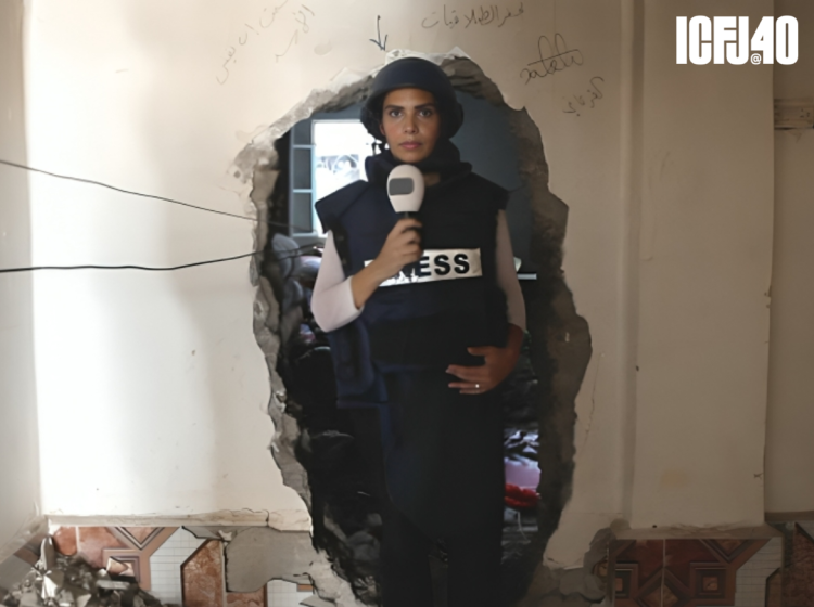 Hadeel Arja stands in the ruins of a building. She's wearing a helmet and a vest that reads "PRESS" while holding a microphone.