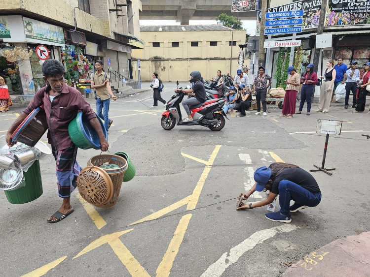 A journalist from the SLPI workshop crouches on a busy Sri Lankan street. She angles her phone up to capture a photo of a man walking with many baskets.