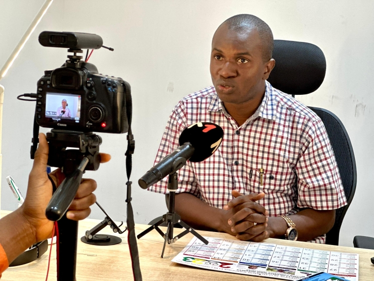 A man sits at a table, speaking into a microphone and camera.