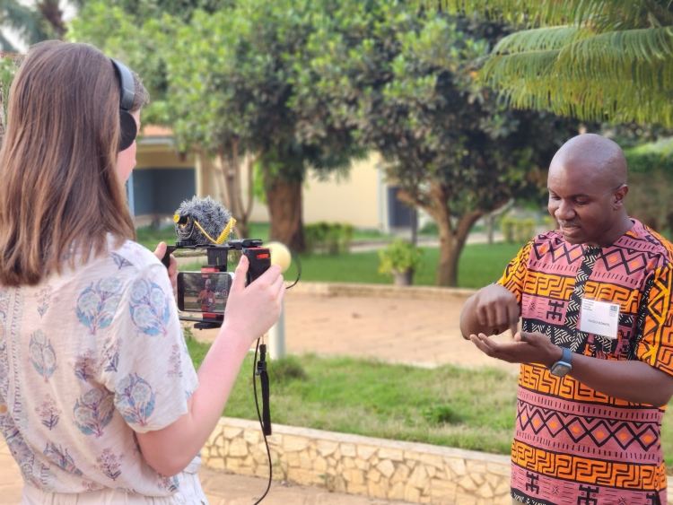 In the foreground, Facely Konaté is being interviewed by a person with a camera and microphone. They're standing outside with trees and large sidewalks in the background.