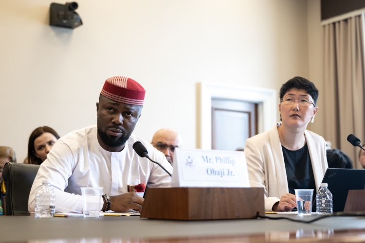 Journalist Philip Obaji speaks into a microphone at a table, before a name card bearing his name and alongside another witness.