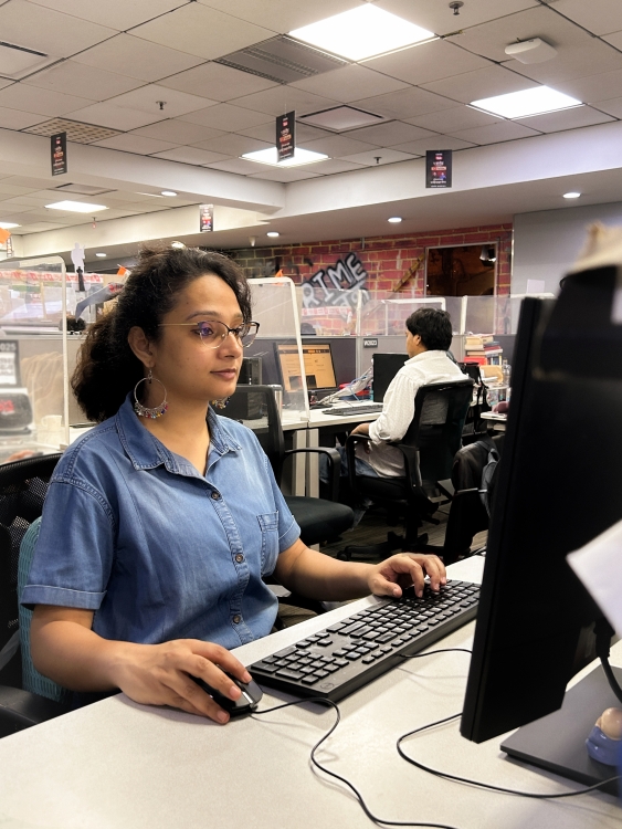 A photo of Jyoti in a newsroom, sitting at her desk and using a computer. 