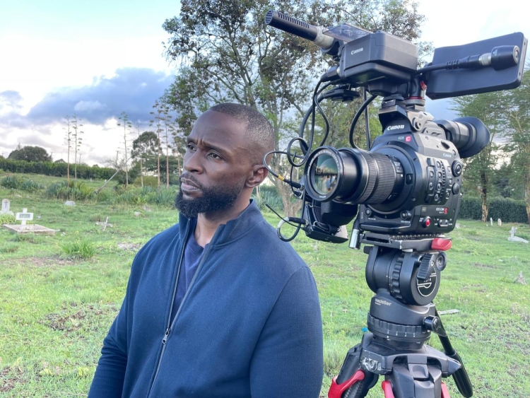 A photo of John-Allan Namu in a field, standing next to a camera on a tripod.