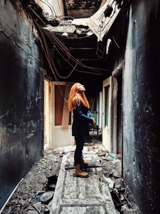 A photo of Valeriya Yegoshyna in a ruined building. She's standing in what looks to have once been a hallway, staring up at a hole in the ceiling.