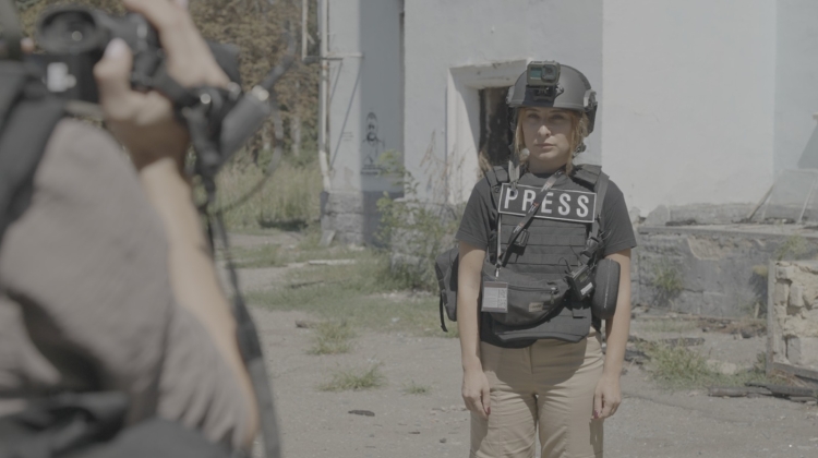 A photo of Valeriya Yegoshyna giving a report for a camera. She's wearing a helmet and a bulletproof vest with the word "PRESS" on it.