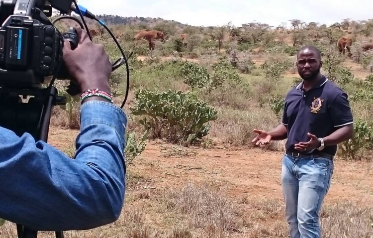 A photo of John-Allan Namu outside in a field, talking to a camera. 