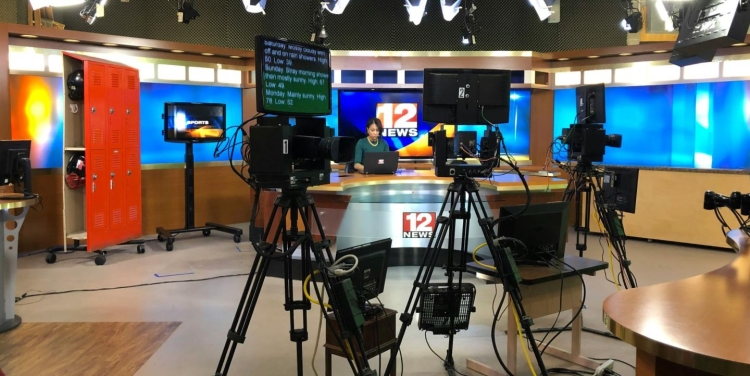 A photo of a broadcast newsroom, with teleprompters and cameras in the foreground and an anchor and anchor desk in the background.