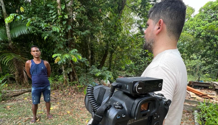 A photo of Daniel standing next to a camera, interviewing a man standing in the camera frame. They're in a forest.