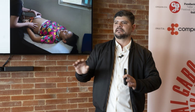 A photo of Daniel Nardin speaking in front of a TV screen. The screen displays an image of a woman receiving an ultrasound.